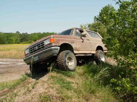 1988 Ford Bronco - A brown Ford Bronco navigates a grassy hill with its front-left tire lifted.