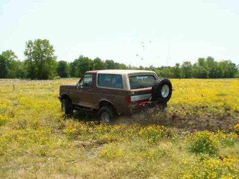 1988 Ford Bronco - A Ford Bronco kicks up mud as it drives through a field of yellow flowers under a clear sky.