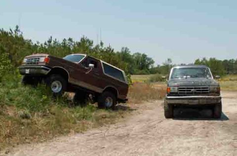 1988 Ford Bronco - Two Ford trucks are parked on a dirt road with one Bronco on a grassy incline.
