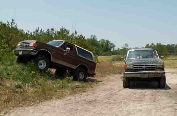 Brown 1987 Ford Bronco on grassy incline next to dark Ford pickup truck on dirt road