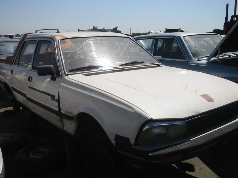 White Peugeot 505 sedan in a junkyard with yellow markings on windshield and missing front wheel