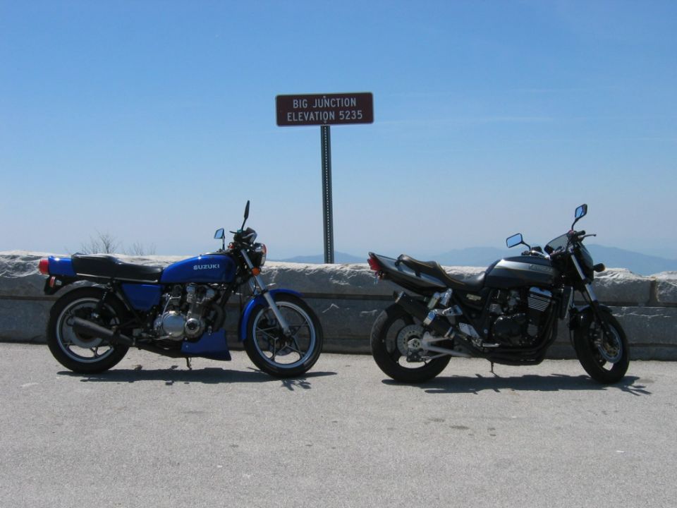 Blue Suzuki motorcycle and a dark-colored motorcycle parked side-by-side on a paved surface with a stone wall and a sign reading "BIG JUNCTION ELEVATION 5235" in the background