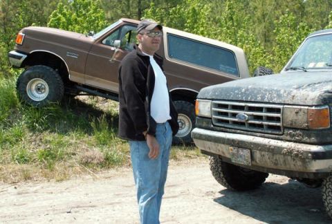 1988 Ford Bronco - Two Ford trucks, a brown Bronco and a black pickup, are parked outdoors on a dirt path with trees in the background.