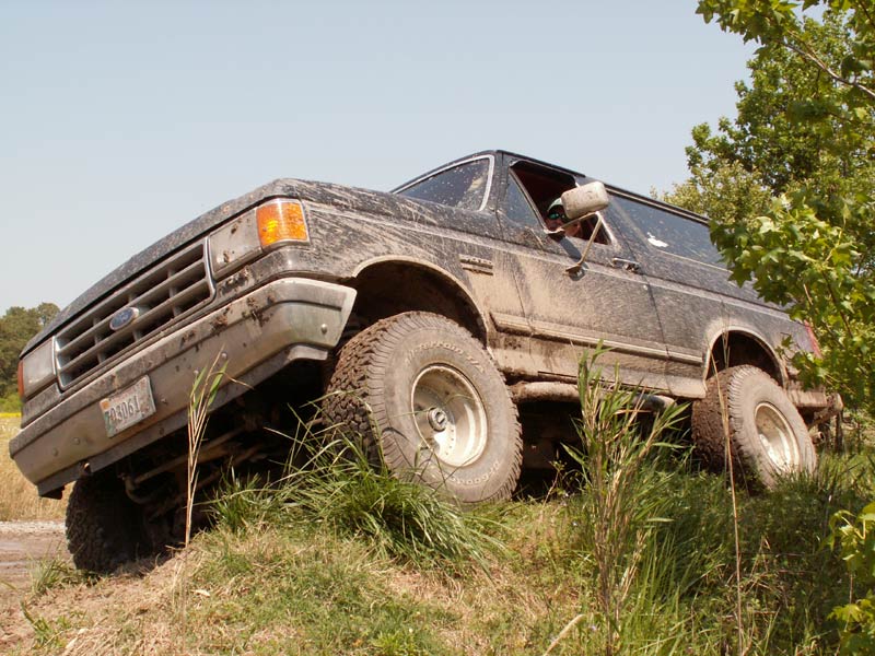 Mud-covered 1987 Ford Bronco on a grassy incline, front 3/4 view