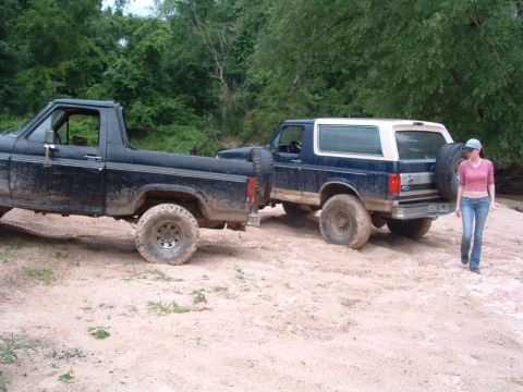 1993 Ford Bronco - Two off-road vehicles are parked on a sandy trail surrounded by trees, with a woman walking away from the Bronco.