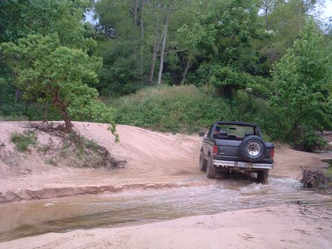 1993 Ford Bronco - A black 1993 Ford Bronco navigates a shallow stream on a dirt path surrounded by trees.
