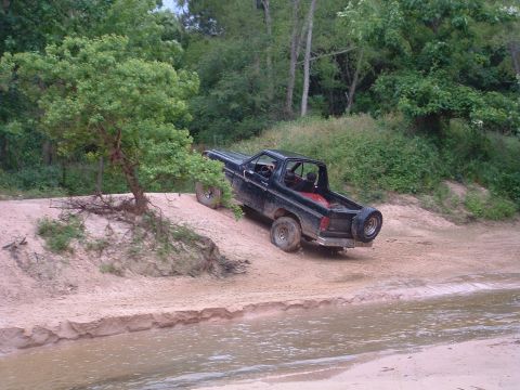 1993 Ford Bronco - This SUV is navigating a sandy incline near a body of water, with trees and foliage in the background.