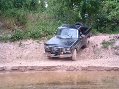 1993 Ford Bronco - Black Ford Bronco navigating a dirt path near water, spare tire mounted on the roof.