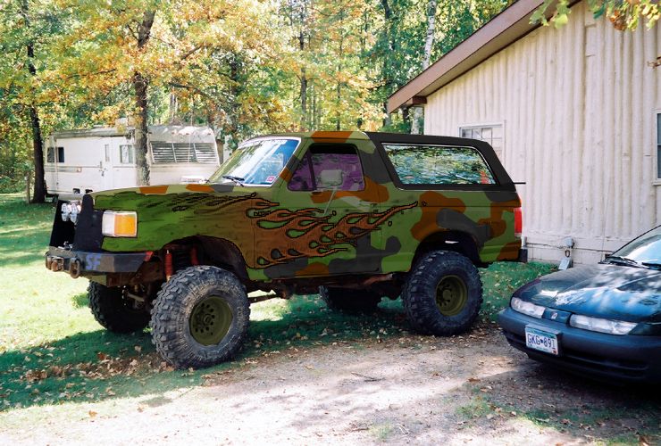 1993 Ford Bronco with camouflage paint and flame decals, parked next to a house
