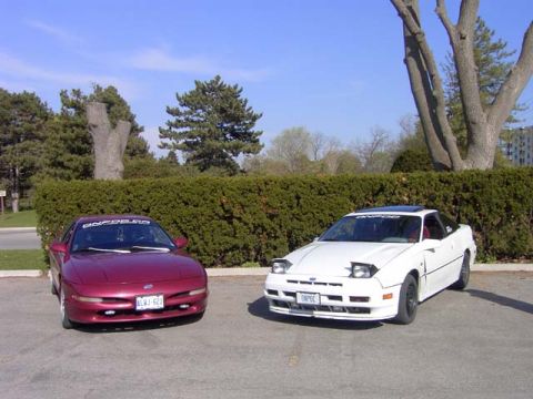 1989 Ford Probe - Two Ford Probes are parked in a lot with a hedge and trees in the background.