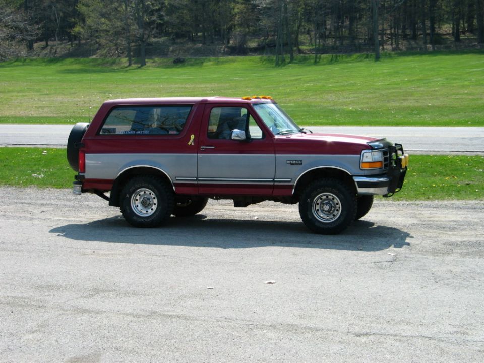 Maroon and silver Ford Bronco parked on gravel lot. Spare tire on back