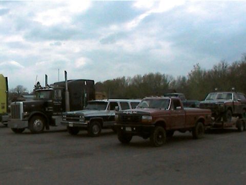 1977 Ford F-150 - A red pickup truck is parked alongside other vehicles, including a large semi-truck and a trailer.