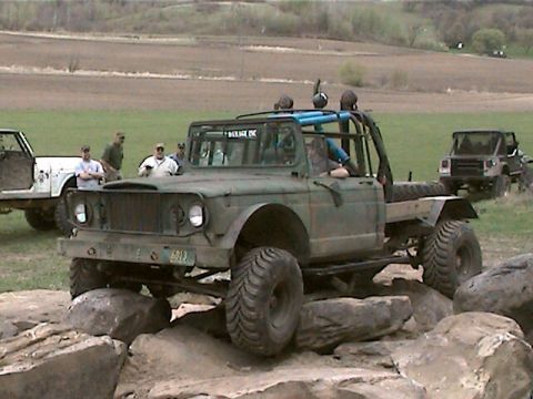 1977 Ford F-150 - An olive green off-road truck with a roll cage is shown navigating a rocky terrain, with other vehicles and people in the background.