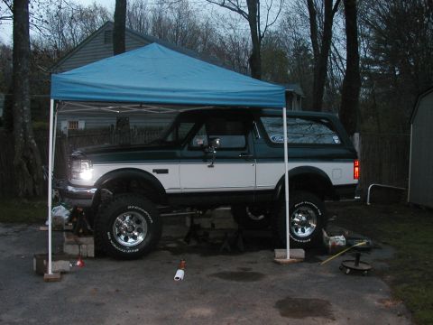 1994 Ford Bronco - A two-tone green and white 1994 Ford Bronco is parked under a blue canopy, elevated on jack stands.