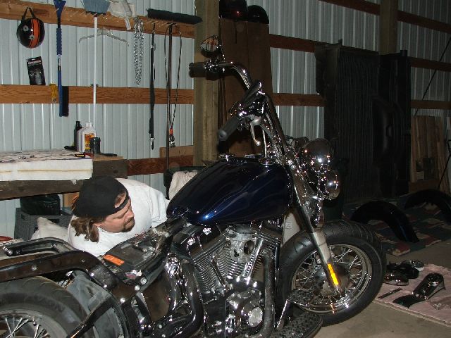 Man working on the chrome V-twin of a dark blue Harley-Davidson Softail in a metal shop