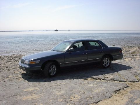 1985 Mercury Grand Marquis - A dark sedan is parked on a rocky shore next to the water under a clear sky.