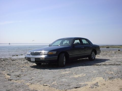 1985 Mercury Grand Marquis - A dark sedan is parked on a rocky beach with the ocean in the background under a clear blue sky.