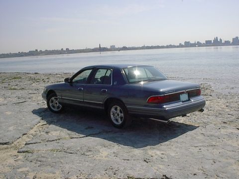 1985 Mercury Grand Marquis - A dark blue Mercury Grand Marquis is parked on a rocky shoreline with a city skyline in the distance.