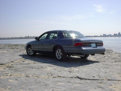 1985 Mercury Grand Marquis - This dark blue sedan is parked on a rocky shore with a city skyline in the distance.