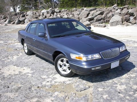 1985 Mercury Grand Marquis - This blue sedan is parked on a rocky surface with trees and houses in the background.