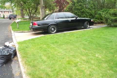 1999 Ford Crown Victoria - Side profile of a black Ford Crown Victoria sedan parked nose-up on a residential concrete driveway with a green lawn in the foreground and…