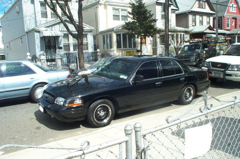 1999 Ford Crown Victoria - Black Ford Crown Victoria sedan parked at the curb on a residential street of clapboard houses, seen from the driver's-side front three-qua…