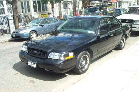 1999 Ford Crown Victoria - A black Ford Crown Victoria Police Interceptor parked at a city curb, front three-quarter view showing a blacked-out grille, black steel wh…