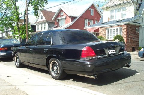 1999 Ford Crown Victoria - Rear three-quarter view of a black Ford Crown Victoria parked at the curb on a residential street, wearing tinted glass, steel police-style…