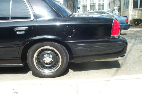 1999 Ford Crown Victoria - Rear quarter close-up of a black Ford Crown Victoria parked at a curb, showing the fuel filler door, red tail lamp, chrome bumper strip and…