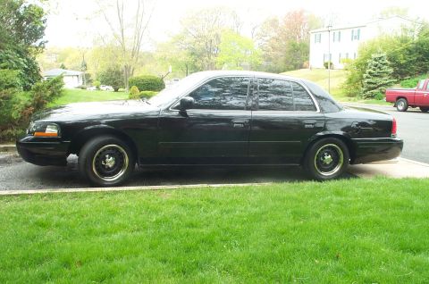 1999 Ford Crown Victoria - Full driver-side profile of a black Ford Crown Victoria sedan on a concrete driveway with a lawn in the foreground and suburban houses and…