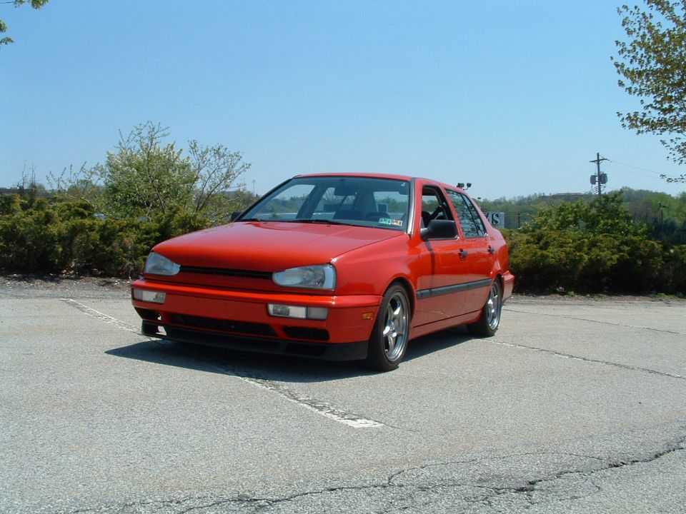 Red sedan parked on asphalt, front 3/4 view. Aftermarket wheels visible
