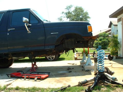 1995 Ford Bronco - A blue Ford Bronco is undergoing front-end suspension work, with parts laid out on the ground.
