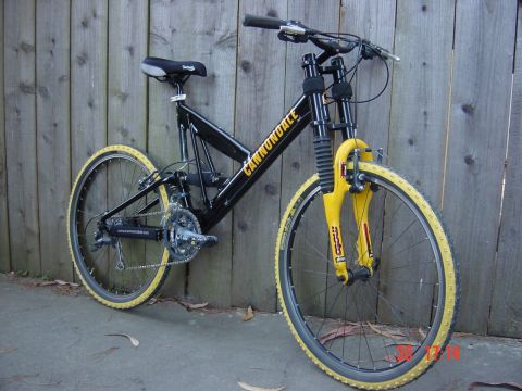 1990 Ford Bronco - A black Cannondale bicycle with yellow tires and front suspension is parked against a weathered wooden fence.