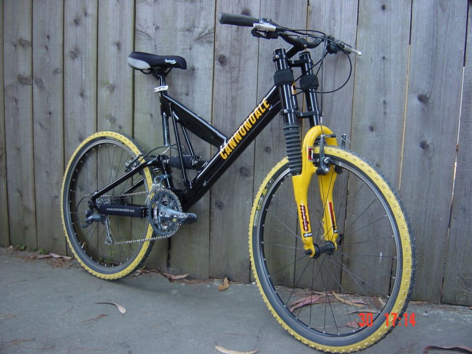 Black Cannondale bicycle with yellow tires and front suspension, leaning against a wooden fence