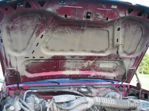 1995 Ford Bronco - The engine bay of a dusty red vehicle, with the hood open, revealing engine parts and the underside of the hood.