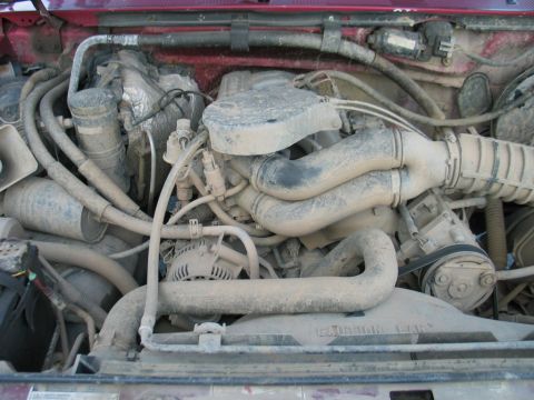 1995 Ford Bronco - The engine bay of a vehicle, covered in dust, shows the intake manifold, hoses, and alternator.