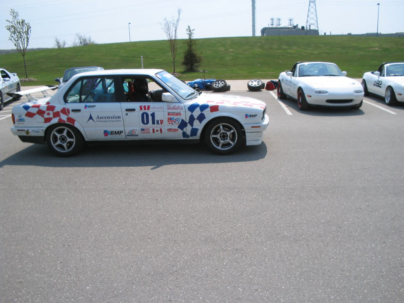 White sedan race car #01 KP with red/blue checkered graphics parked near two white Miatas in a paddock lot
