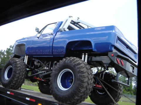 1989 Ford Bronco - A blue monster truck with oversized tires and custom suspension is loaded onto a flatbed trailer.