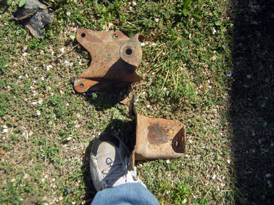 1995 Ford Bronco - Two rusty metal brackets lie on the ground next to a person's shoe.