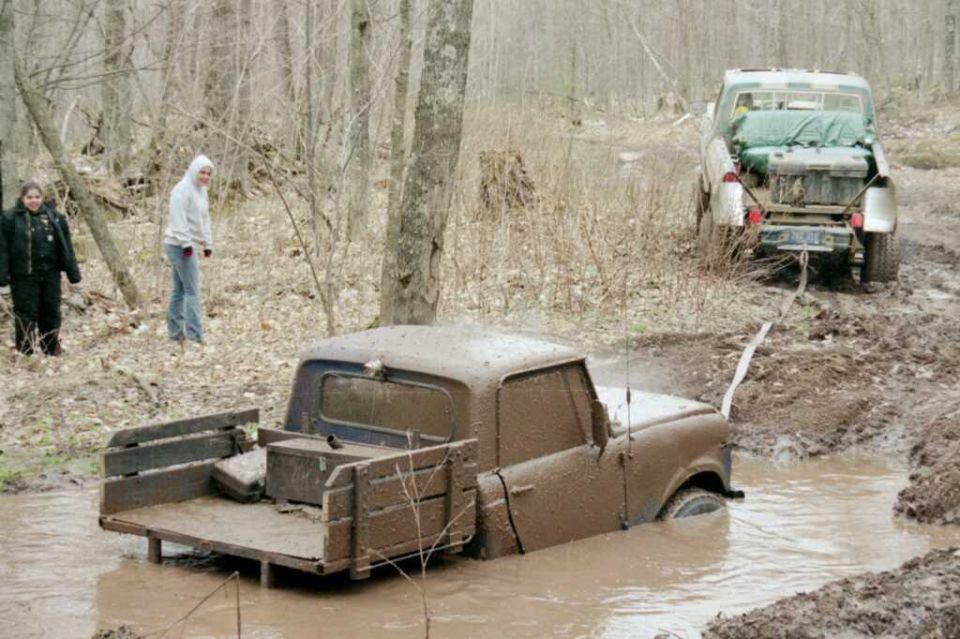 Mud-covered Scout pickup stuck in a deep mud pit with a larger truck behind it in the woods