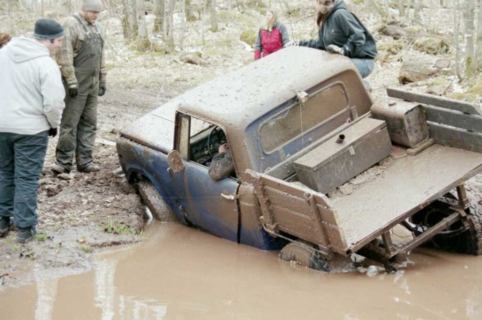 Blue off-road truck stuck and sinking in a deep muddy water pit with onlookers