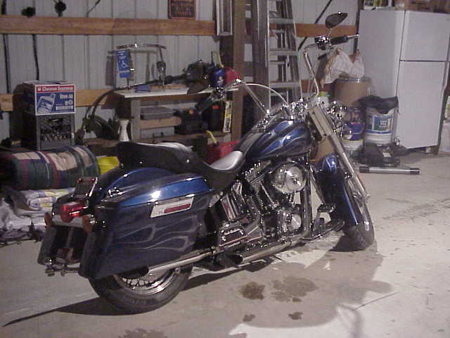 Dark blue ghost-flame Harley-Davidson Softail with saddlebags parked in a shop, rear 3/4 view