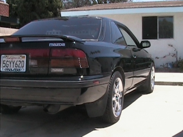 Black Mazda turbo coupe from the rear three-quarter in a driveway on polished aftermarket wheels