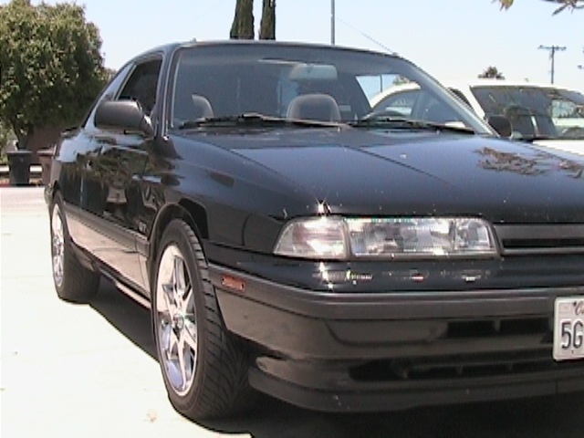 Black first-gen Mazda MX-6 coupe with chrome wheels, three-quarter front view in a sunny lot