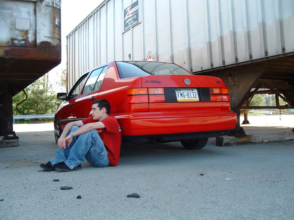 Red 1994 Volkswagen Jetta sedan parked under a trailer, man sitting on ground next to it