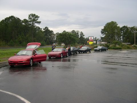 1997 Ford Probe - A row of hatchback coupes lined up on a wet parking lot under overcast skies, led by a red car with its hood, hatch and driver's door open…