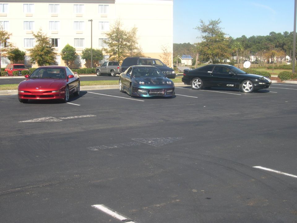 Three sports cars parked in a lot: a red Ford Probe, a teal modified car with hood open, and a black car with 'GTX' badge