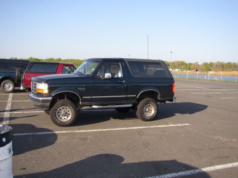 1995 Ford Bronco - A dark green Ford Bronco is parked in an asphalt lot next to other vehicles, with a body of water in the background.