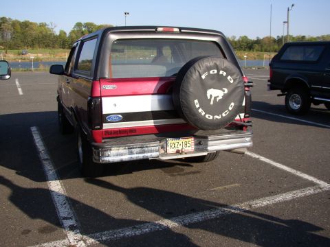 1995 Ford Bronco - A red and black Ford Bronco is parked in a lot, showing its rear with a spare tire cover and license plate.
