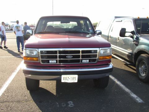 1995 Ford Bronco - A maroon Ford Bronco is parked in a lot, with another truck visible to its right and people in the background.